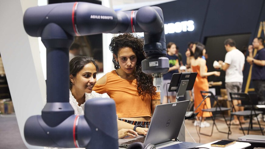 Two people are working at a trade fair stand on a laptop in front of which a robot arm can be seen.