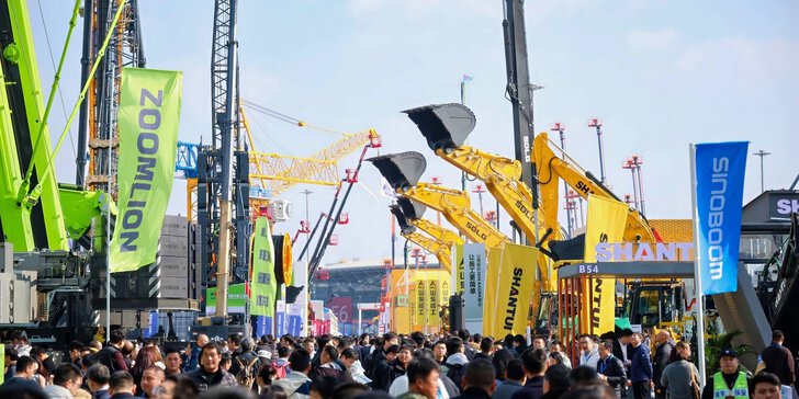 A crowd at an outdoor construction equipment exhibition, with cranes, excavators, and company banners in view.