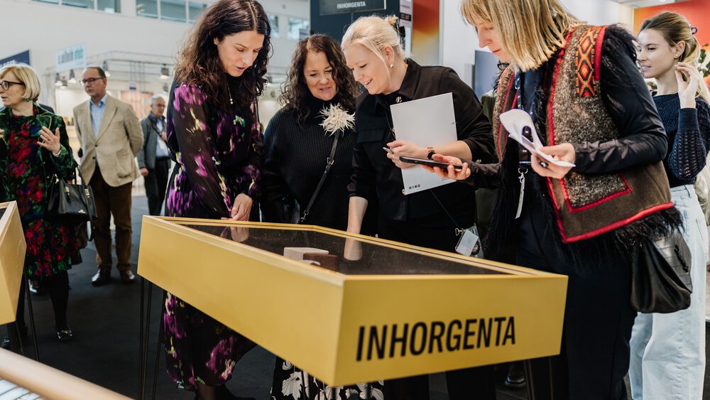 A group of women examine a display case at the INHORGENTA trade fair, while others watch nearby.