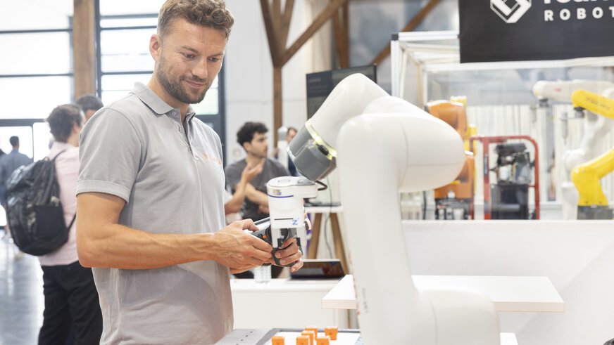 A man tests the remote control of a robotic arm with gripping tool at a trade fair stand.