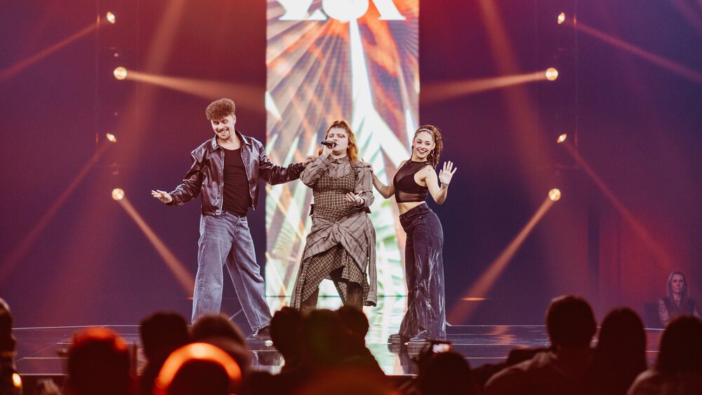 A man and two women stand on a stage and perform. Above their heads hangs a screen with the word 
