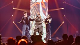 A man and two women stand on a stage and perform. Above their heads hangs a screen with the word 