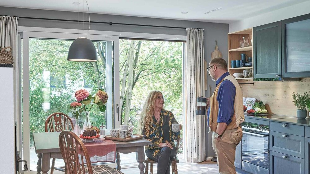 Ein Mann und eine Frau unterhalten sich in einer gemütlich modern eingerichteten Wohnküche mit Blick auf einen üppigen Garten. Ein Hund sitzt zwischen Ihnen.