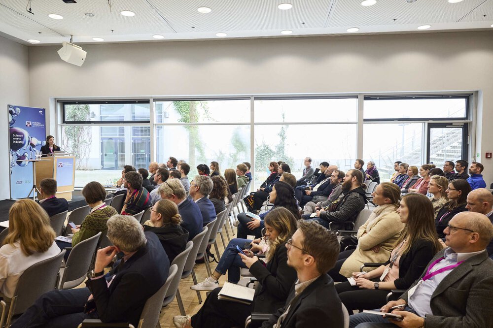 People sit in a conference room and listen to a speaker giving a presentation at a podium near large windows.