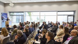 People sit in a conference room and listen to a speaker giving a presentation at a podium near large windows.