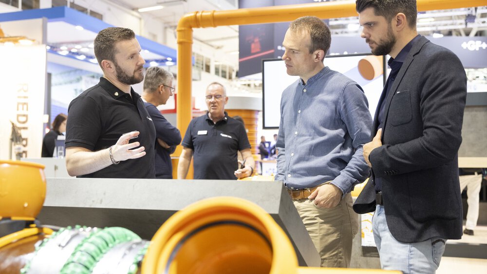 Ein Mann erklärt zwei weiteren Männern in Businesskleidung etwas an einem Stand auf der IFAT Messe in München. Im Vorder- und Hintergrund sind Rohre zu sehen.