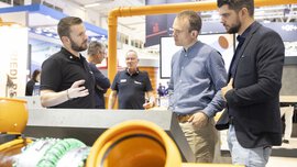 Ein Mann erklärt zwei weiteren Männern in Businesskleidung etwas an einem Stand auf der IFAT Messe in München. Im Vorder- und Hintergrund sind Rohre zu sehen.