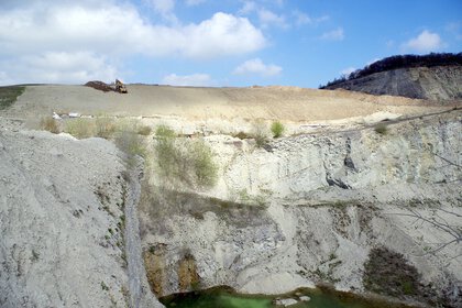 A construction vehicle is working on a large, rocky hill. At the foot of the hill is a small, green pond