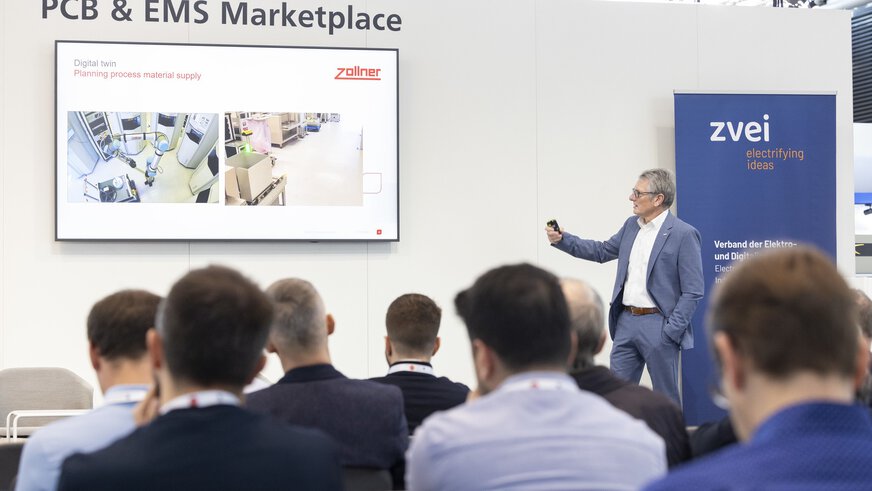 Ein Mann im Anzug steht vor einem Publikum und hält eine Präsentation zum Thema „Digital twin – Planning process material supply“.