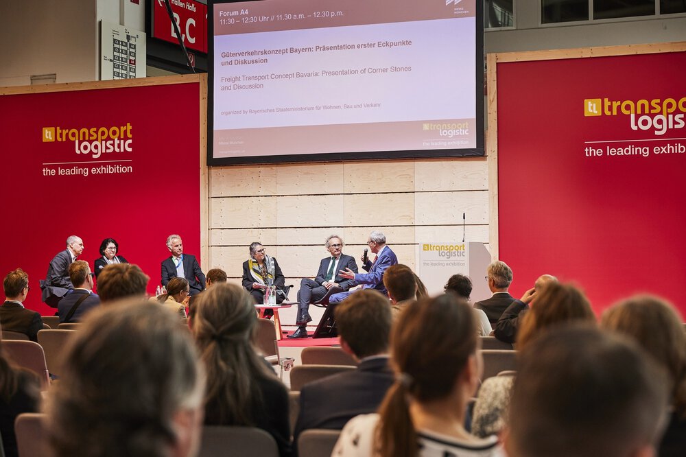 The panelists discuss on the stage of a transportation logistics exhibition while the audience sits and screens are behind them.