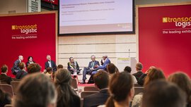 The panelists discuss on the stage of a transportation logistics exhibition while the audience sits and screens are behind them.