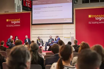 The panelists discuss on the stage of a transportation logistics exhibition while the audience sits and screens are behind them.