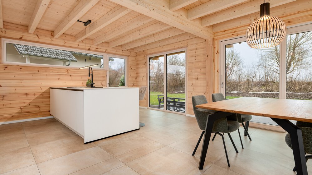 Kücheninterieur aus Holz mit Kochinsel, Esstisch, Stühlen, Hängelampe und großen Fenstern mit Blick auf einen Garten.
