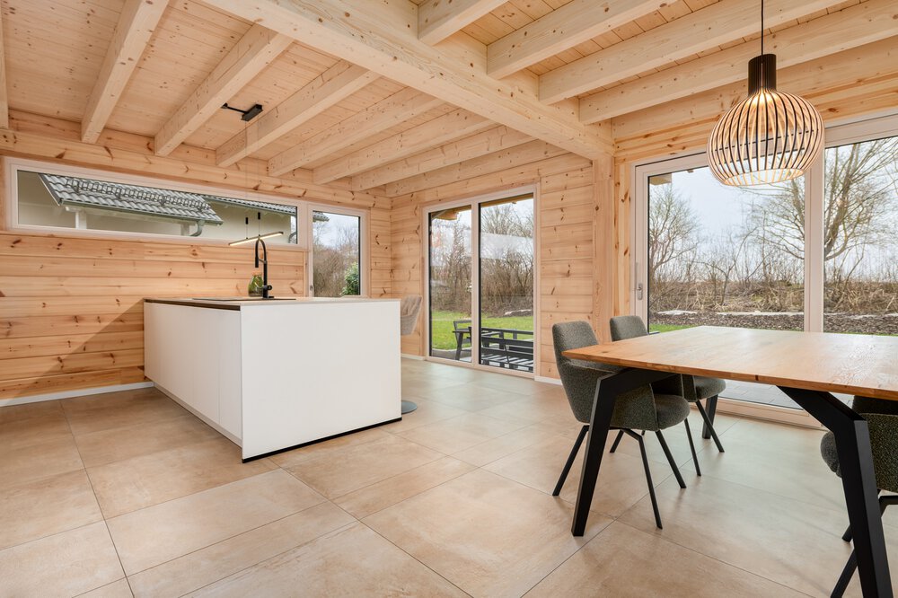 Kücheninterieur aus Holz mit Kochinsel, Esstisch, Stühlen, Hängelampe und großen Fenstern mit Blick auf einen Garten.