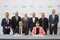 Eight people in suits pose for a group photo; two of them sit at the front and sign contracts at a table.