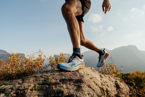 Die durchtrainierten braungebrannten Beine eines Mannes in hellblauen Sportschuhen mit orangener Sohle, die über einen Erdhügel joggen.