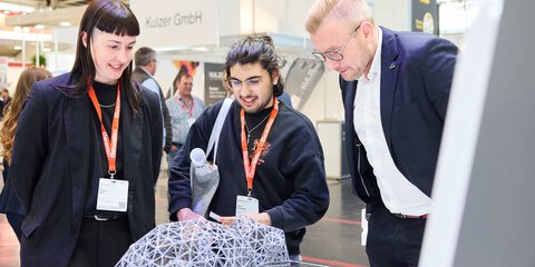 Three trade fair visitors examine a delicate ceramic 3D exhibit together.