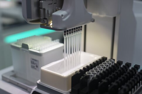 Acht Pipetten an einer Vorrichtung angebracht, die automatisiert Probenröhrchen befüllen, hängen über einen kleinen Schale