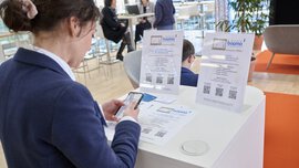 A woman at the bauma Mediendialog 2025 uses her smartphone to vote for the bauma Innovation Award 2025. In the background, other participants engage in conversations in a modern lounge setting.