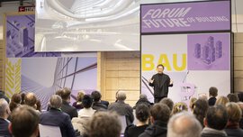 The speaker presents architectural designs to an audience at a building forum, which are displayed on a screen.
