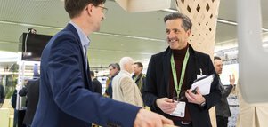 Two men in business attire smile at each other while standing at a stand at BAU.