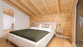 Schlafzimmer mit Holzwänden und -decke, einem Doppelbett mit grüner Decke, Kissen und Beistelltischchen mit Nachttischlampen.