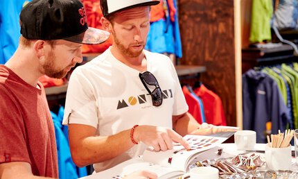 Zwei Männer stehen an einem Tisch in einem Ausstellungsraum und betrachten die Artikel in einem aufgeschlagenen Katalog.