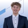 Ein lächelnder Mann mit lockigen rot-braunen Haaren trägt ein weißes Oberhemd und ein dunkelblaues Jackett darüber.