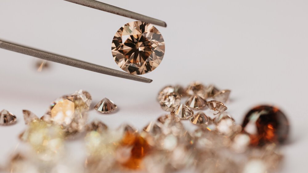 A pair of tweezers is holding a cut diamond aloft. Beneath it lie further cut diamonds in similar colours on a light-coloured background.