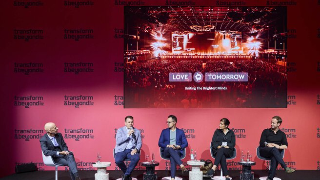 Eine Podiumsdiskussion auf der Bühne „Transform & Beyond“ der EXPO REAL in München. Fünf Expertinnen und Experten sitzen auf der Bühne und diskutieren, während einer der Teilnehmenden spricht und ein Mikrofon hält. Hinter ihnen ist eine große Leinwand mit einem Bild einer Veranstaltung und dem Schriftzug „Love Tomorrow – Uniting the Brightest Minds“ zu sehen. Die Bühne ist in kräftigem Pink gestaltet und vermittelt eine moderne, dynamische Atmosphäre.