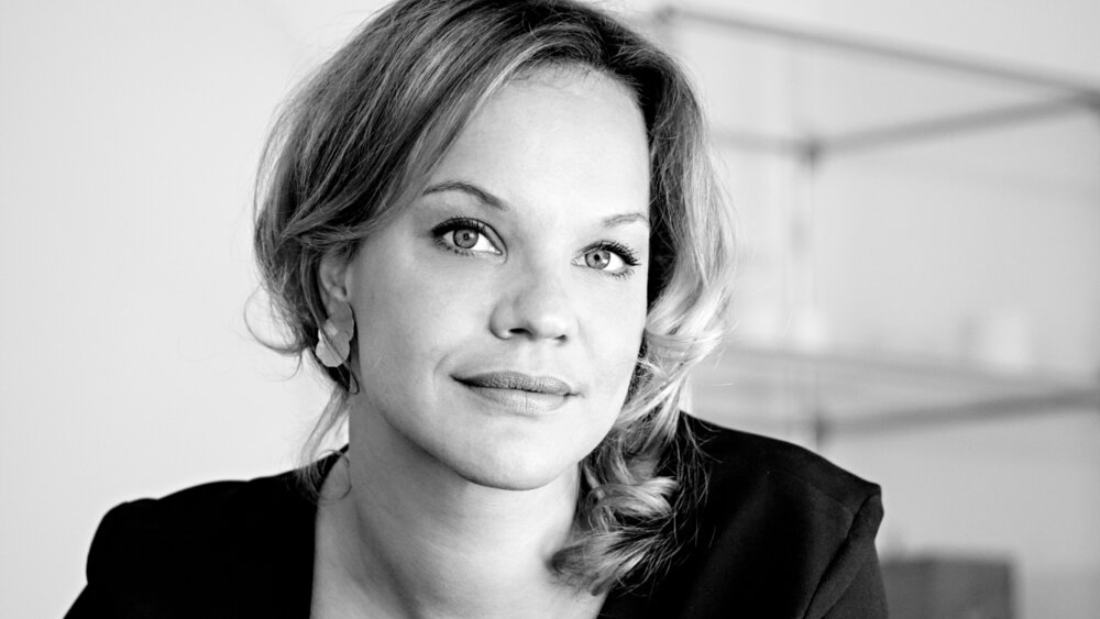 A smiling woman with shoulder-length curly hair. She is wearing a dark top, earrings and a ring on her finger.
