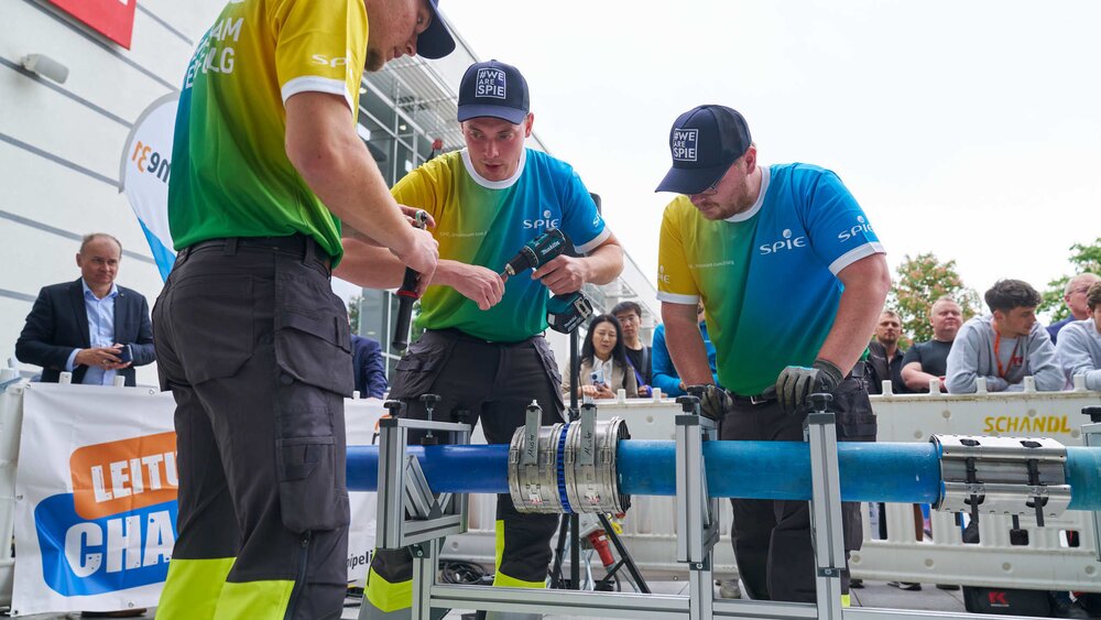 Three technicians in team shirts work together on a pipe joint during a competition at the trade fair's outdoor area.