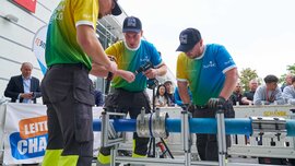 Three technicians in team shirts work together on a pipe joint during a competition at the trade fair's outdoor area.