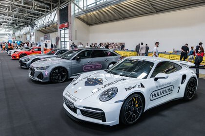 Auf einer Automobilausstellung sind im Innenbereich mehrere Sportwagen ausgestellt, darunter ein weißer Porsche mit Sponsorenaufklebern. Die Besucher schlendern umher und betrachten die Fahrzeuge.