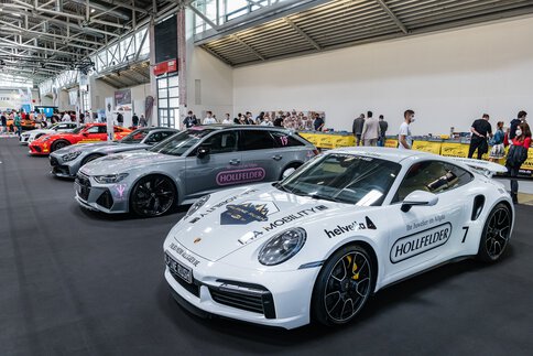 Auf einer Automobilausstellung sind im Innenbereich mehrere Sportwagen ausgestellt, darunter ein weißer Porsche mit Sponsorenaufklebern. Die Besucher schlendern umher und betrachten die Fahrzeuge.