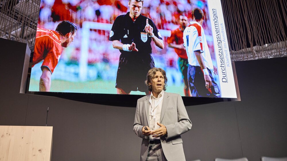 Der ehemalige Fußballschiedsrichter Urs Meier hält einen Vortrag auf der EXPO REAL in München. Hinter ihm ist auf einer großen Leinwand ein Bild eines Fußballschiedsrichters während eines Spiels zu sehen, daneben der Schriftzug „Durchsetzungsvermögen – Klarheit im Ausdruck“. Der Vortragende trägt einen hellgrauen Anzug und spricht engagiert vor dem Publikum.
