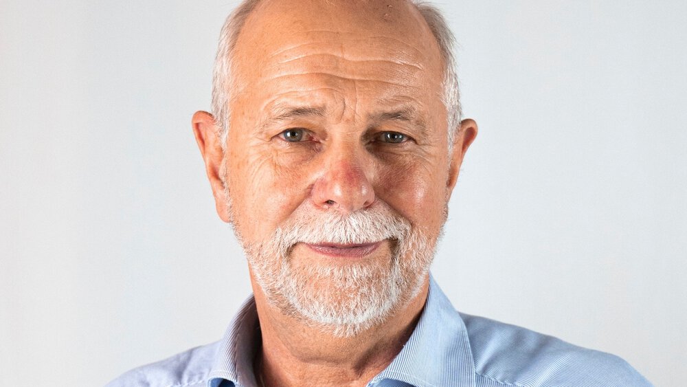 A smiling man with grey hair and beard in a blue shirt.
