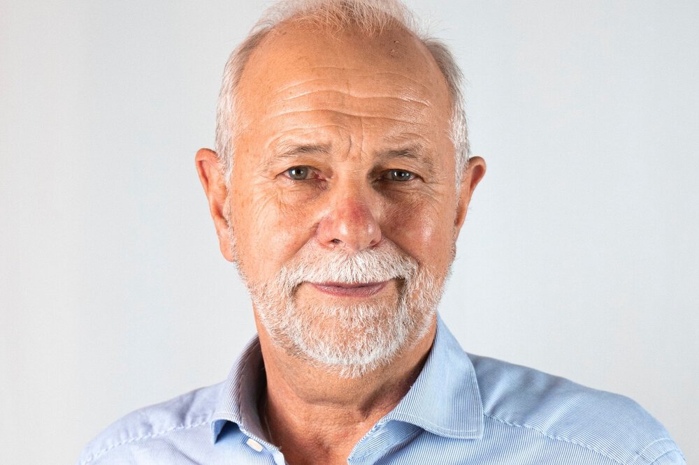 A smiling man with grey hair and beard in a blue shirt.