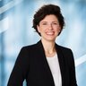 Eine lächelnde Frau mit kurzen schwarzen Haaren, in einem schwarzen Blazer über einer weißen Bluse.