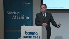 Ein Mann im Anzug präsentiert auf dem bauma Forum 2022 neben einem Banner mit der Aufschrift 