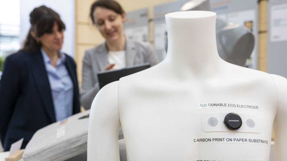 The white torso of a mannequin with a technical device and several lines of text stuck to it. Two women are conversing in the blurred background.