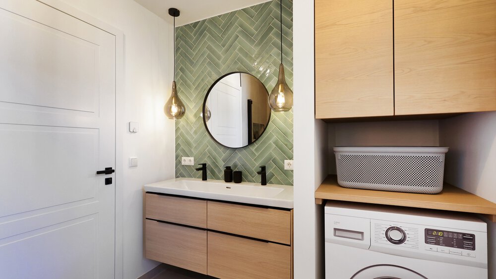 Modernes Badezimmer mit Holzschränken, Doppelwaschbecken, rundem Spiegel, grün gefliester Wand und Waschmaschine.