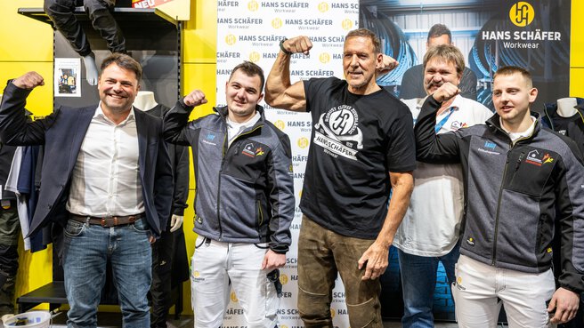 Five men stand in front of a trade fair stand for workwear and pose with Ralf Möller with their right arm raised to show off their muscles.