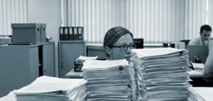  In an office, a woman wearing glasses is sitting at a desk behind tall piles of documents, whilst another person is working in the background.