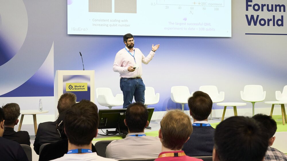 Ein Sprecher präsentiert auf der Bühne der World of Quantum in München vor Publikum, im Hintergrund ist eine Folie zur Quantenforschung zu sehen.