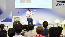 Ein Sprecher präsentiert auf der Bühne der World of Quantum in München vor Publikum, im Hintergrund ist eine Folie zur Quantenforschung zu sehen.