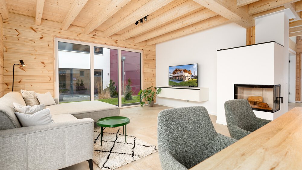 Modernes Wohnzimmer mit hellen Holzwänden, gemütlichen Möbeln, einem an der Wand montierten Fernseher und großen Fenstern mit Blick auf den Garten.