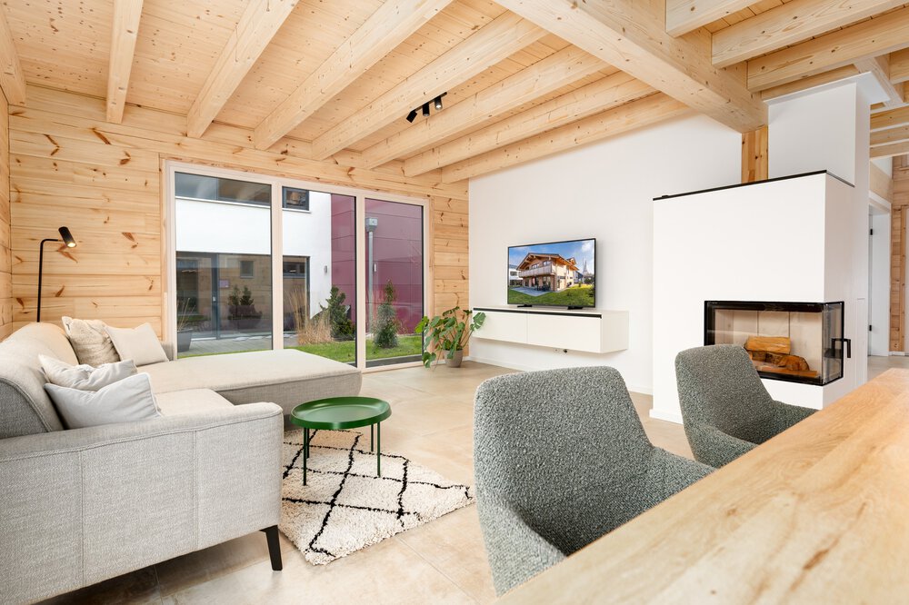 Modernes Wohnzimmer mit hellen Holzwänden, gemütlichen Möbeln, einem an der Wand montierten Fernseher und großen Fenstern mit Blick auf den Garten.