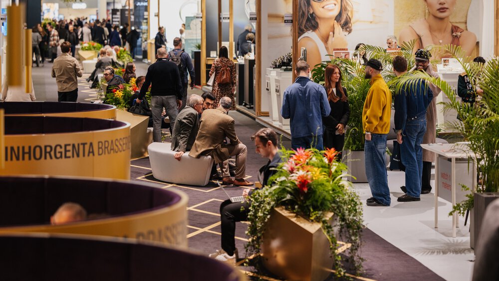 Menschen versammeln sich und interagieren bei einer Indoor-Veranstaltung mit Pflanzen, Sitzgelegenheiten und großen Bildern von Frauen im Hintergrund.