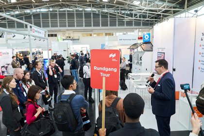 Guided trade fair tour with a group of visitors and a guide speaking into a microphone.